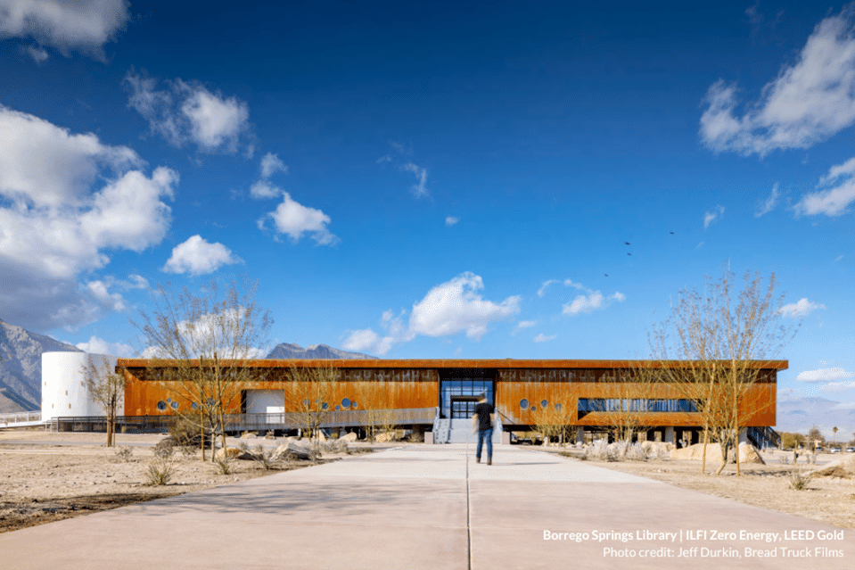 Modern single story building with light wood exterior in the middle of a desert with bright blue skies above. Text reads: "Borrego Springs Library | ILFI Zero Energy, LEED Gold."
