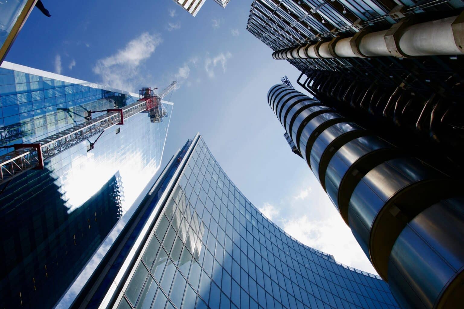 Looking up at modern skyscrapers against a blue sky, with glass facades reflecting sunlight. A crane stretches between the buildings, conveying urban growth.