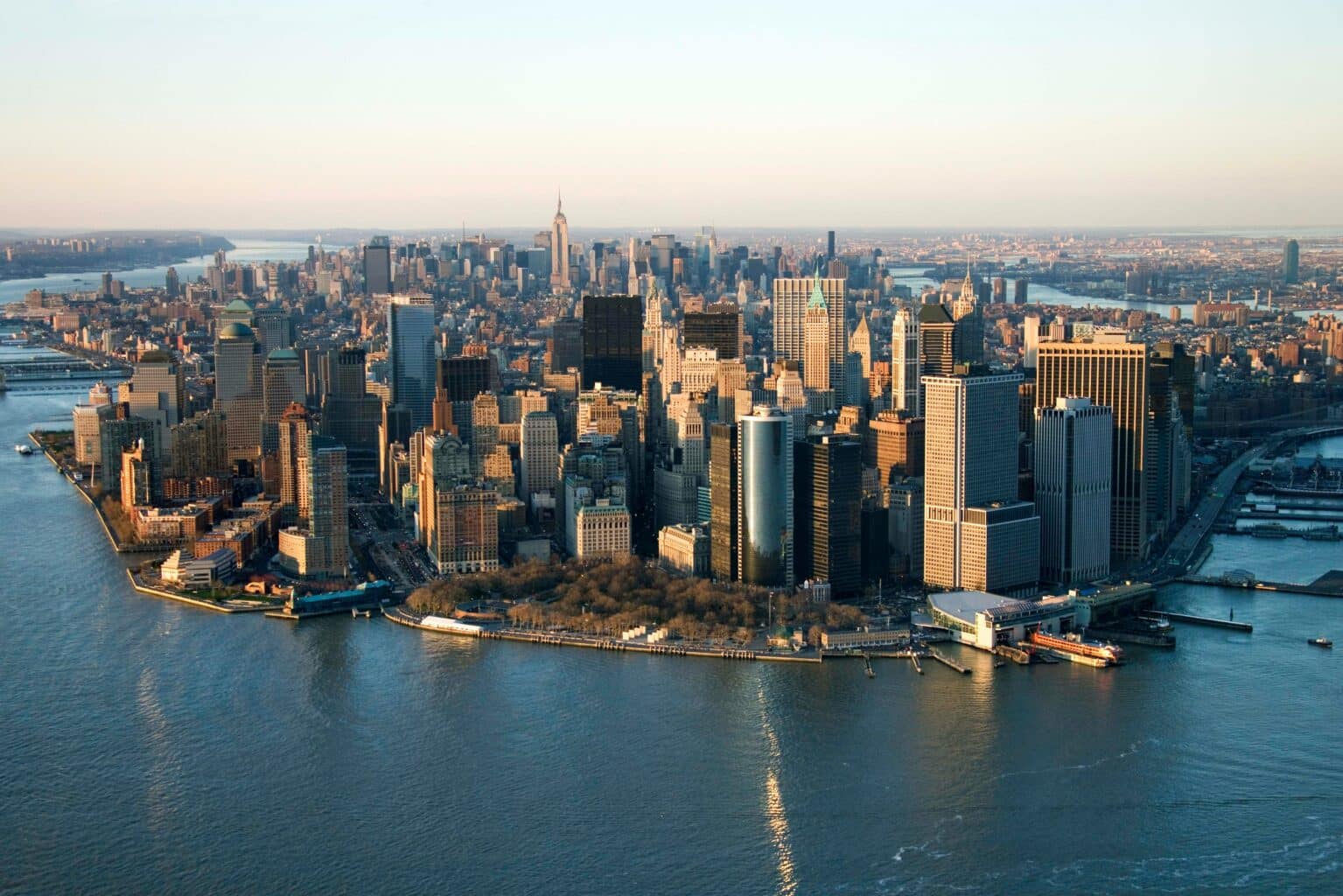 Aerial view of Manhattan at sunset, showcasing skyscrapers with reflective windows surrounded by rivers. The skyline conveys an urban and vibrant atmosphere.
