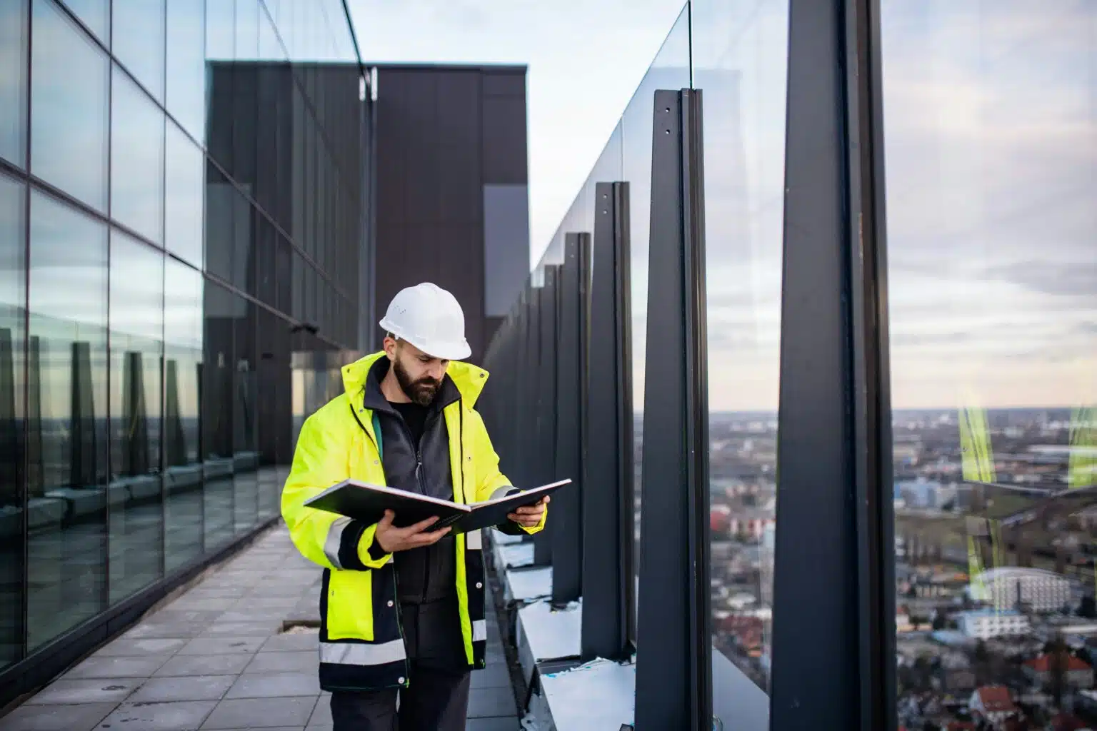 A construction worker in a yellow safety jacket and hard hat reviews blueprints on a high-rise rooftop with a glass railing, overlooking a cityscape.